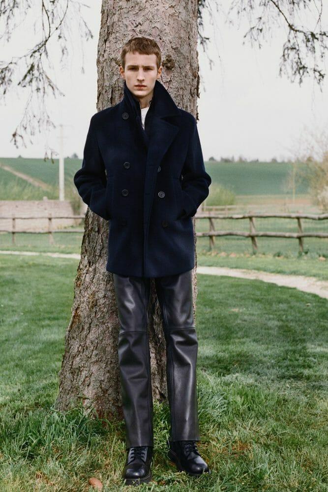 Modelo apoyado en un árbol con pantalones y abrigo oscuros de Sandro