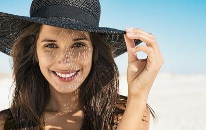 Rostro de una mujer joven sonriendo en la playa con un sombrero negro protegiendo su piel