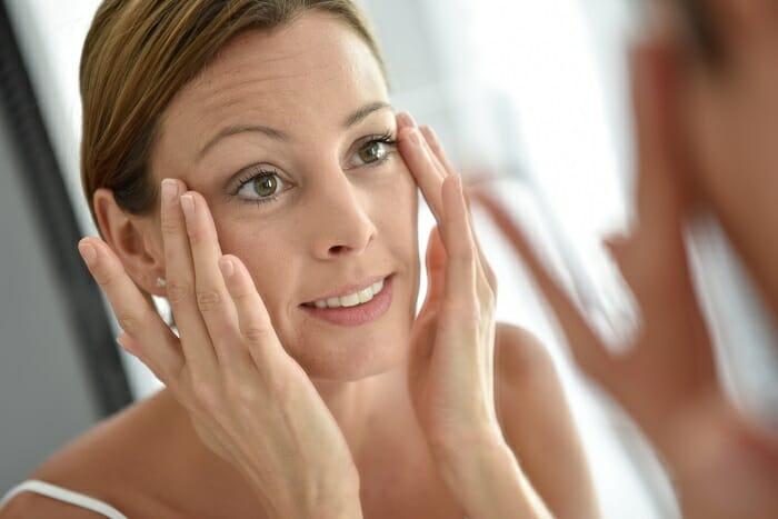 Mujer mirándose al espejo y echándose con sus manos una crema facial en la cara