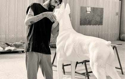 Fotografía en blanco y negro del artista David Morago con una escultura en forma de perro Doberman