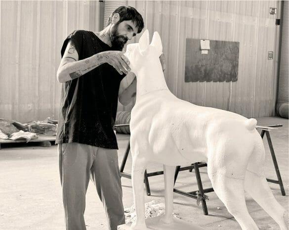 Fotografía en blanco y negro del artista David Morago con una escultura en forma de perro Doberman