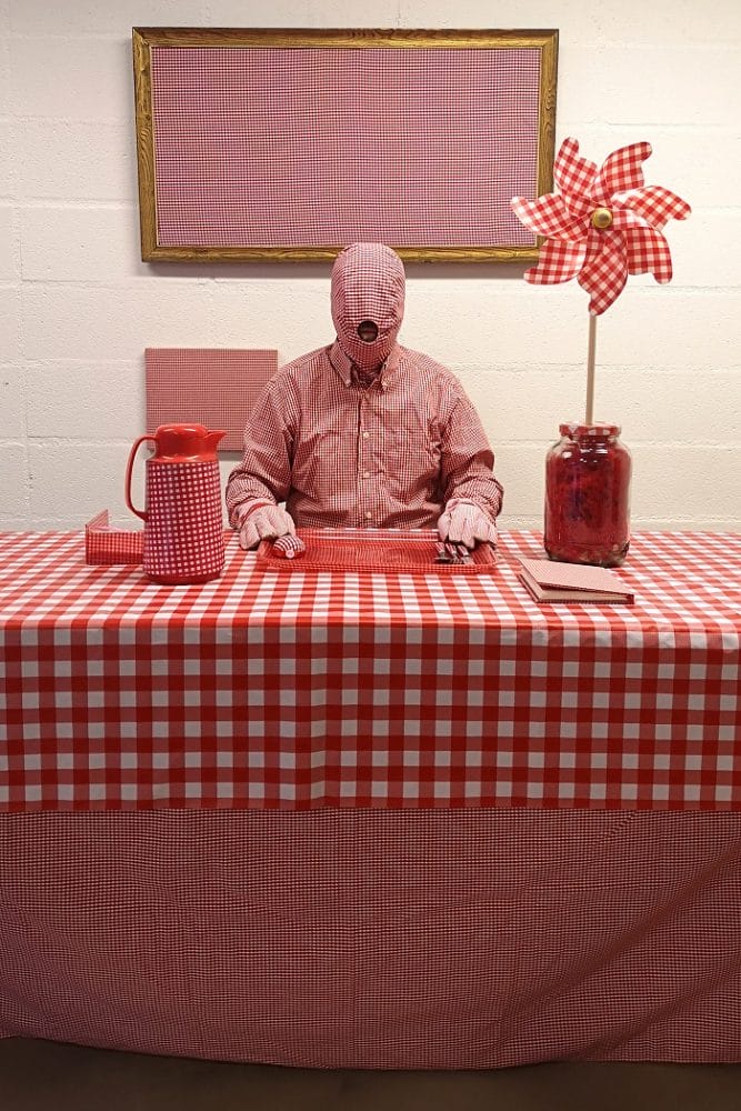 Fotografía de un señor encapuchado en una mesa con mantel de cuadros blancos y rojos con jarra y florero a juego